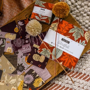 May include: A wooden tray displays fabric squares with autumn-themed patterns and two pom-pom decorations. The fabric squares feature fall leaves and floral designs in shades of orange, brown, and green. The text "Windfall" is visible on the fabric packaging.