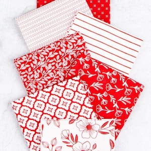 May include: A stack of folded fabric squares in various patterns. The fabrics feature red and white designs, including floral, geometric, and striped patterns. The fabrics are arranged on a white surface.
