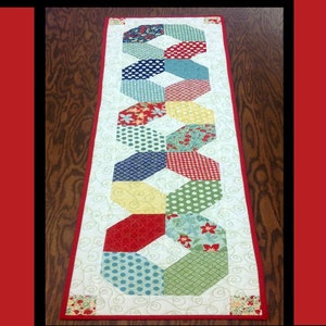 May include: A colorful table runner with a geometric pattern of hexagons in red, blue, green, yellow, and white fabrics. The runner is called "Penny Candy" and is designed by Tricia Lynn Maloney for Orphan Quilt Designs.