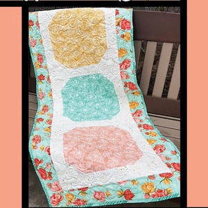 May include: A quilted table runner with the text "Window Boxes." It features three large, irregularly shaped blocks in yellow, teal, and pink, each with a floral pattern. The edges are bordered with a floral print.