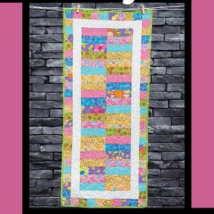 May include: A colorful quilt with the word "Bubblegum" at the top. The quilt features a patchwork design with various colors, including pink, blue, yellow, and green. It is framed with a white border and a green edge. The quilt is hanging against a dark brick wall.