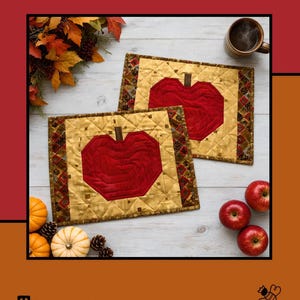 May include: Two rectangular quilted placemats featuring a central red apple motif on a gold background with a dark patterned border. The text Apples Mellow, Tamara Cook for Villa Rosa Designs is visible on the product packaging along with autumn decor.