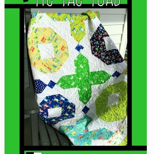 May include: A green, blue, yellow, and white quilt with a tic-tac-toe pattern and a frog theme. The quilt is folded and sitting on a black chair. The quilt is titled "Tic Tac Toad" and is by Tricia Lynn Maloney, Orphan Quilt Designs for Villa Rosa Designs.