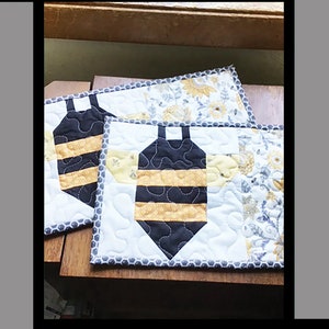May include: Two quilted bee-shaped snack mats in black, white, and yellow fabric. The mats are on a wooden surface. The text "Buzzy Bee Snack Mat" is at the top of the image. The text "Tricia Lynn Maloney Orphan Quilt Designs for Villa Rosa Designs" is at the bottom of the image.