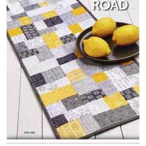 May include: A rectangular patchwork quilt with yellow, gray, and white squares. The quilt is labeled "Mini BRICK ROAD". Three lemons sit on a dark plate. The image also includes the text "ATKINSON DESIGNS".