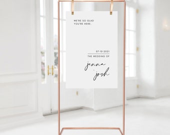 May include: A copper-colored metal wedding sign with a white paper insert. The sign reads "WE'RE SO GLAD YOU'RE HERE." and "07-10-2021 THE WEDDING OF jenna + josh" in black script. The sign is displayed indoors.