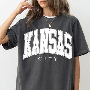 Peut inclure: Un t-shirt gris anthracite &agrave; manches courtes avec le texte KANSAS CITY en grandes lettres majuscules blanches sur la poitrine. Le t-shirt pr&eacute;sente une encolure ronde et une coupe d&eacute;contract&eacute;e, avec le mot KANSAS arqu&eacute; au-dessus de CITY.