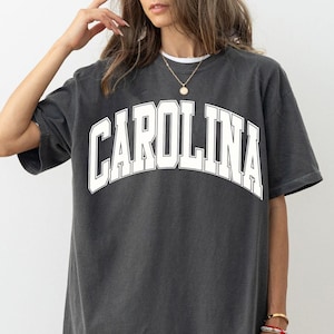 May include: A charcoal grey oversized t-shirt featuring the word CAROLINA printed in large white block letters across the chest in an arched design. The garment has a crew neckline, short sleeves, and a relaxed fit with a faded, washed-out texture.