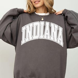 May include: A charcoal gray crewneck sweatshirt featuring the word INDIANA printed in large white block letters with a thin white outline across the chest. The garment has long sleeves with ribbed cuffs and a ribbed hem, providing a relaxed fit.