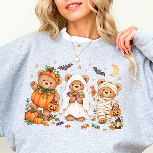 May include: A sport grey crewneck sweatshirt featuring a graphic of three brown teddy bears dressed for Halloween. One bear wears a pumpkin costume, one is a ghost, and one is a mummy. They are surrounded by bats, stars, a moon, and candy.