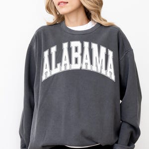 May include: A charcoal grey crewneck sweatshirt featuring the word ALABAMA printed in large white block letters across the chest in an arched design. The garment has long sleeves with ribbed cuffs and a ribbed neckline, worn over a white shirt.