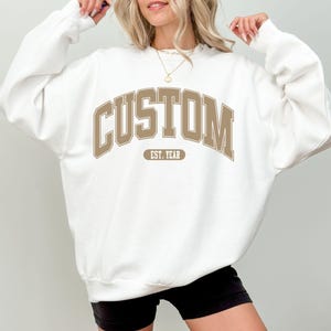 May include: White sweatshirt with the word "CUSTOM" in a brown collegiate font across the chest. Below the word "CUSTOM" is the text "EST. YEAR" in a smaller brown font. The sweatshirt is a casual style.