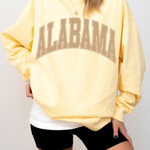 May include: A pale yellow long-sleeve crewneck sweatshirt featuring the word ALABAMA printed in a large, arched, tan-colored collegiate font across the chest. The garment has a relaxed fit with ribbed cuffs and a ribbed hemline.