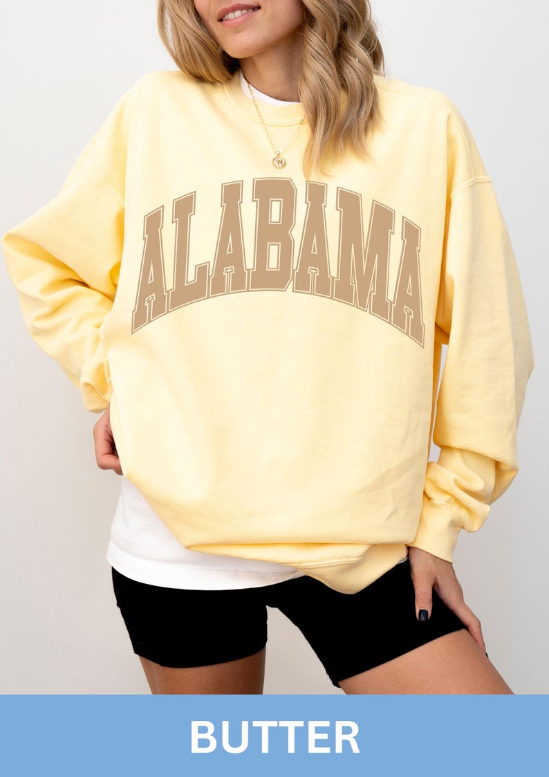 May include: A pale yellow long-sleeve crewneck sweatshirt featuring the word ALABAMA printed in a large, arched, tan-colored collegiate font across the chest. The garment has a relaxed fit with ribbed cuffs and a ribbed hemline.