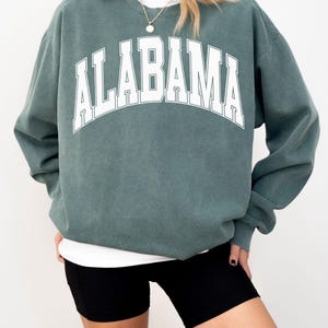 May include: A blue spruce crewneck sweatshirt featuring the word ALABAMA printed in large white capital letters across the chest in an arched font. The garment has long sleeves, a ribbed neckline, and a relaxed fit, layered over a white shirt.