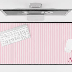 May include: A rectangular desk mat featuring a light pink and white vertical striped pattern. The mat has a black border and the letters AF printed in a dark pink serif font in the bottom right corner. A white keyboard and mouse sit on the mat.
