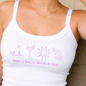 May include: White tank top with a scoop neck and thin straps. The shirt features a purple graphic of a bikini, cocktail, sun, and seashell. Text at the bottom reads "MARY'S BACH/ MIAMI, FL 2026".