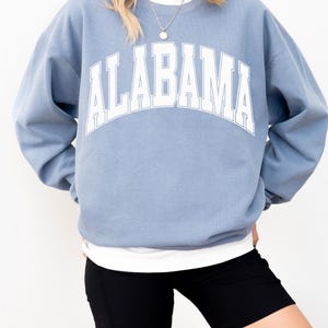 May include: A dusty blue crewneck sweatshirt featuring the word ALABAMA printed in a large, arched white font with a thin blue outline. The long-sleeved pullover has a ribbed collar, cuffs, and hem, worn over a plain white undershirt.
