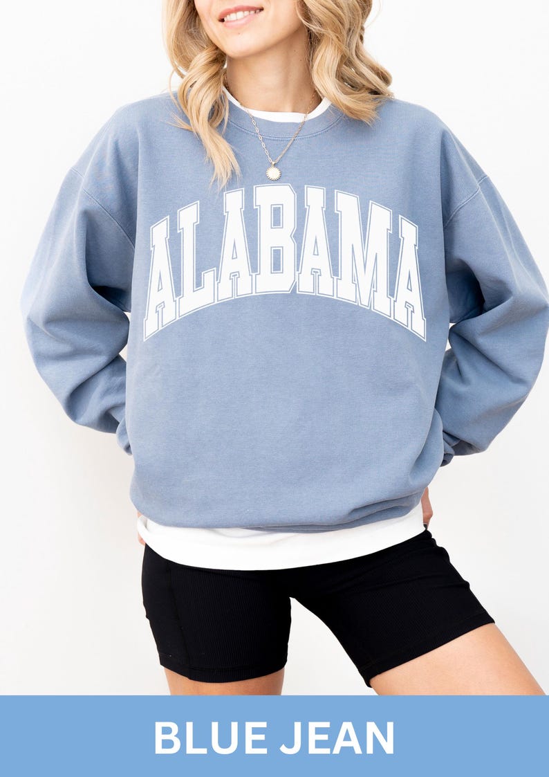 May include: A dusty blue crewneck sweatshirt featuring the word ALABAMA printed in a large, arched white font with a thin blue outline. The long-sleeved pullover has a ribbed collar, cuffs, and hem, worn over a plain white undershirt.