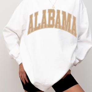 May include: A white crewneck sweatshirt featuring the word ALABAMA printed in a large, arched, tan-colored collegiate font across the chest. The garment has long sleeves with ribbed cuffs and a ribbed hem, providing a relaxed fit.