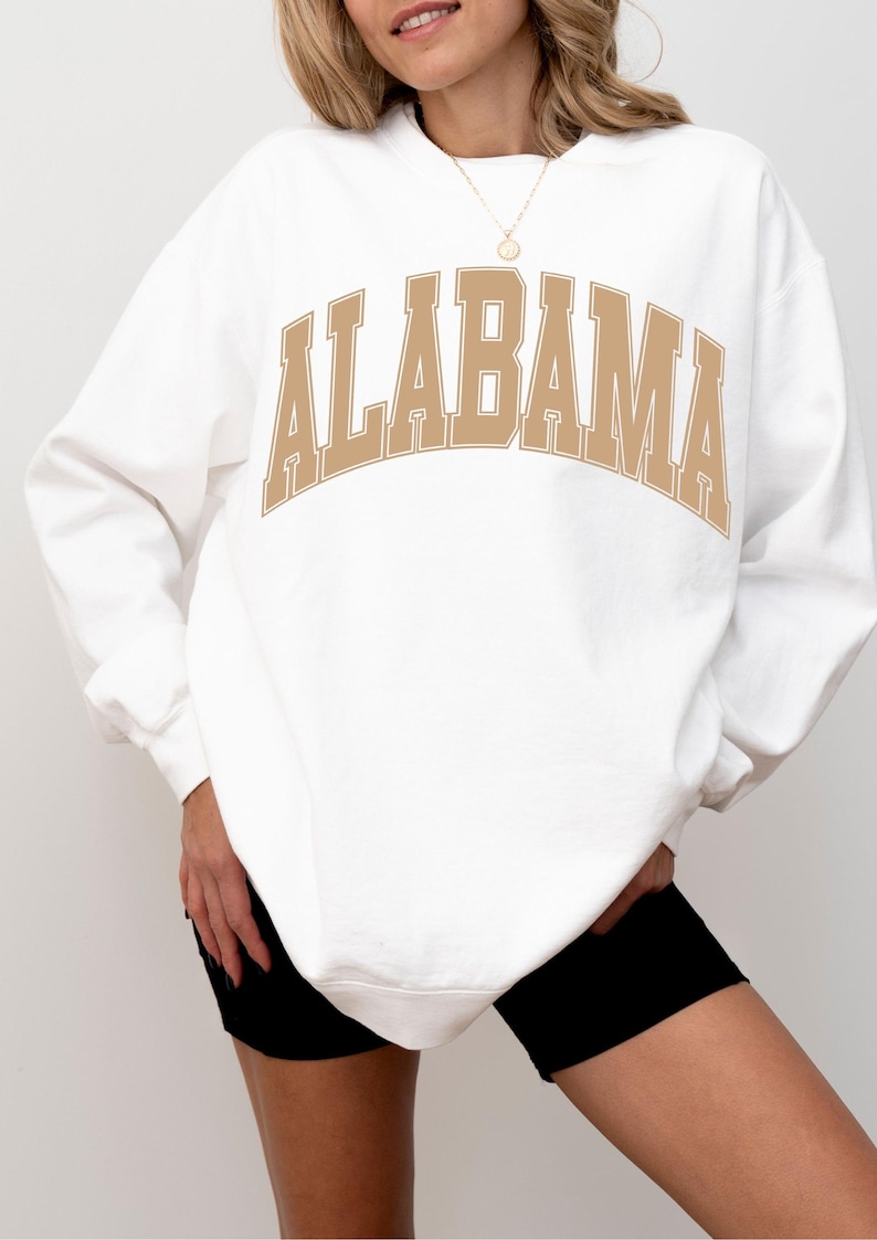 May include: A white crewneck sweatshirt featuring the word ALABAMA printed in a large, arched, tan-colored collegiate font across the chest. The garment has long sleeves with ribbed cuffs and a ribbed hem, providing a relaxed fit.