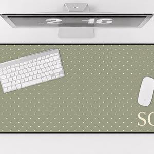 May include: A rectangular desk mat featuring a sage green background with a repeating pattern of small white polka dots. The bottom right corner displays the capital letters SC in a serif font. A white keyboard and mouse rest on the mat.
