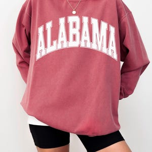 May include: A crimson long-sleeve crewneck sweatshirt featuring the word ALABAMA printed in large white block letters across the chest in an arched design. The garment has a relaxed fit with ribbed cuffs and a ribbed neckline.