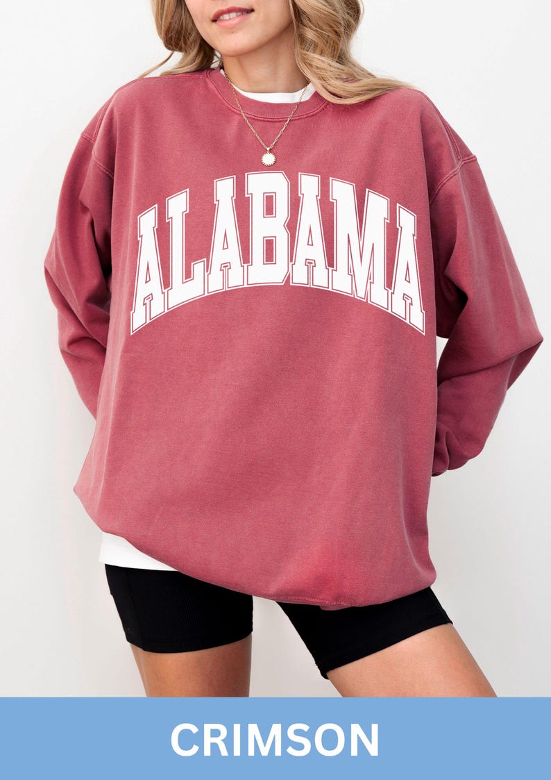 May include: A crimson long-sleeve crewneck sweatshirt featuring the word ALABAMA printed in large white block letters across the chest in an arched design. The garment has a relaxed fit with ribbed cuffs and a ribbed neckline.