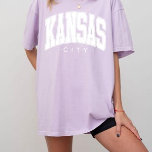 Peut inclure: Un t-shirt oversize violet clair avec le texte KANSAS CITY imprim&eacute; en lettres blanches de style universitaire sur la poitrine. Ce v&ecirc;tement pr&eacute;sente des manches courtes, une encolure ronde et une coupe d&eacute;contract&eacute;e avec une &eacute;criture lisible.
