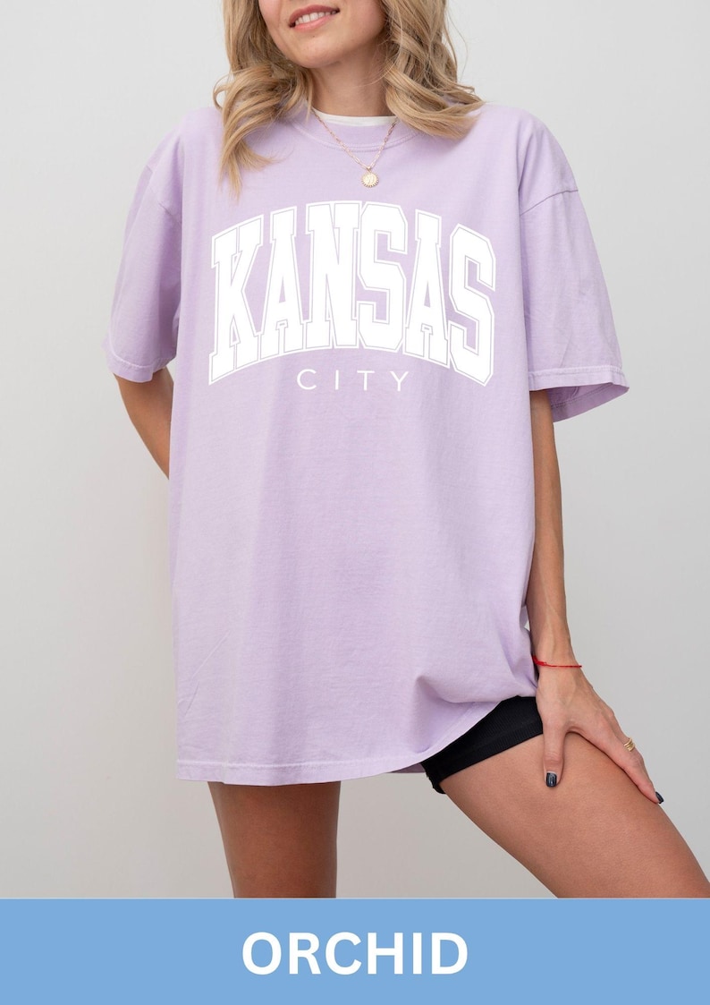 Peut inclure: Un t-shirt oversize violet clair avec le texte KANSAS CITY imprim&eacute; en lettres blanches de style universitaire sur la poitrine. Ce v&ecirc;tement pr&eacute;sente des manches courtes, une encolure ronde et une coupe d&eacute;contract&eacute;e avec une &eacute;criture lisible.