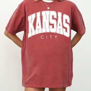 Peut inclure: Un t-shirt &agrave; manches courtes et col rond de couleur rouge cramoisi d&eacute;lav&eacute;. Le devant du v&ecirc;tement pr&eacute;sente le texte KANSAS CITY en grandes lettres capitales blanches et grasses avec un fin contour blanc, centr&eacute; sur la poitrine.