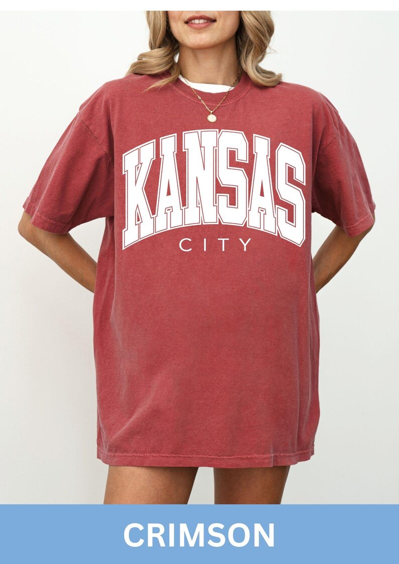 Peut inclure: Un t-shirt &agrave; manches courtes et col rond de couleur rouge cramoisi d&eacute;lav&eacute;. Le devant du v&ecirc;tement pr&eacute;sente le texte KANSAS CITY en grandes lettres capitales blanches et grasses avec un fin contour blanc, centr&eacute; sur la poitrine.