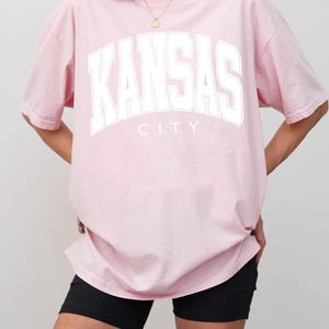 Peut inclure: Un t-shirt rose clair &agrave; manches courtes avec le texte KANSAS CITY imprim&eacute; en grandes lettres blanches sur la poitrine. Le v&ecirc;tement pr&eacute;sente une encolure ronde et une coupe d&eacute;contract&eacute;e, offrant un aspect simple avec sa teinte pastel.