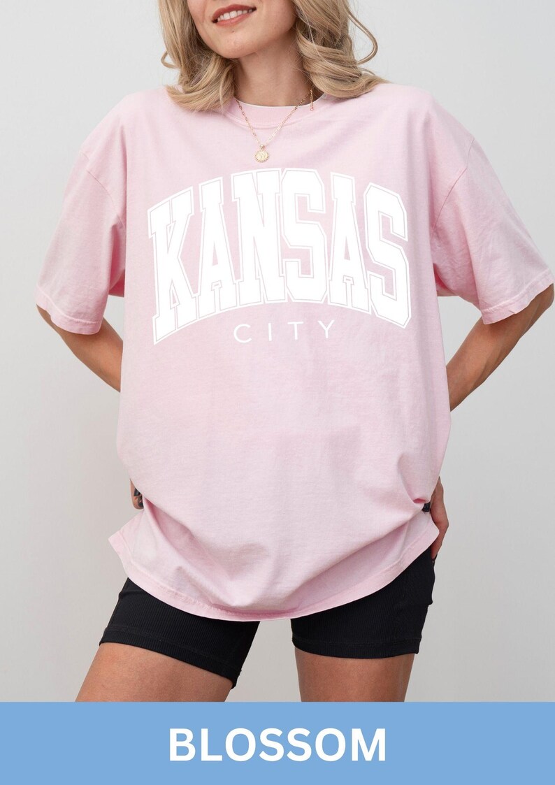 Peut inclure: Un t-shirt rose clair &agrave; manches courtes avec le texte KANSAS CITY imprim&eacute; en grandes lettres blanches sur la poitrine. Le v&ecirc;tement pr&eacute;sente une encolure ronde et une coupe d&eacute;contract&eacute;e, offrant un aspect simple avec sa teinte pastel.