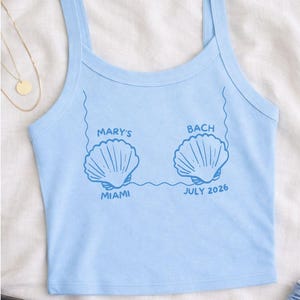 May include: Light blue tank top with a seashell graphic and the text "MARY'S BACH MIAMI JULY 2026". The top has thin straps and a scoop neckline. The design is in a darker shade of blue.
