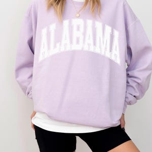 May include: A light purple crewneck sweatshirt featuring the word ALABAMA printed in large white block letters across the chest. The garment has long sleeves and a ribbed collar, worn over a white undershirt with black athletic shorts.