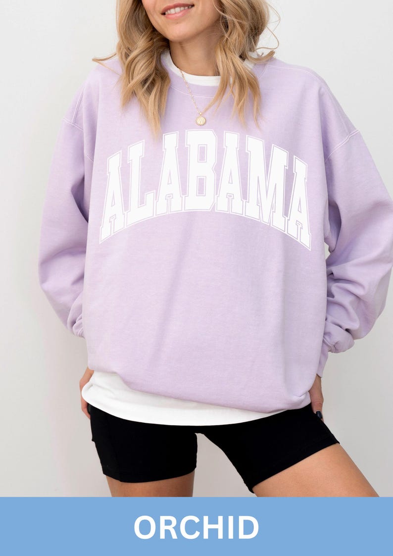 May include: A light purple crewneck sweatshirt featuring the word ALABAMA printed in large white block letters across the chest. The garment has long sleeves and a ribbed collar, worn over a white undershirt with black athletic shorts.