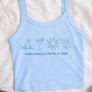 May include: Light blue tank top with thin straps. The front features blue line art of a bikini, cocktail, sun, and seashell. Text below reads "MARY'S BACH/ MIAMI, FL 2026".