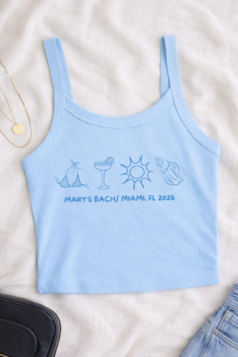 May include: Light blue tank top with thin straps. The front features blue line art of a bikini, cocktail, sun, and seashell. Text below reads "MARY'S BACH/ MIAMI, FL 2026".