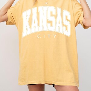 Peut inclure: Un t-shirt &agrave; manches courtes jaune moutarde avec le texte KANSAS CITY en grandes lettres majuscules blanches sur la poitrine. Le t-shirt pr&eacute;sente une encolure ronde et une coupe ample et d&eacute;contract&eacute;e avec une finition de couleur unie.