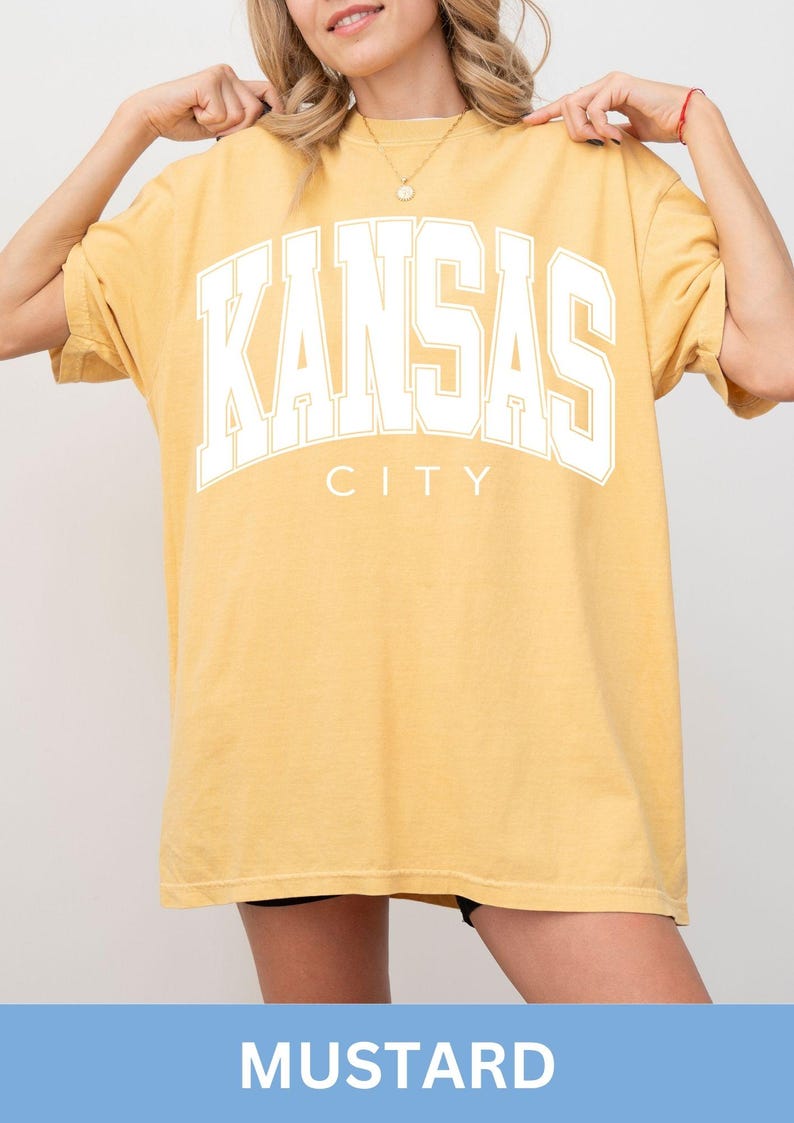 Peut inclure: Un t-shirt &agrave; manches courtes jaune moutarde avec le texte KANSAS CITY en grandes lettres majuscules blanches sur la poitrine. Le t-shirt pr&eacute;sente une encolure ronde et une coupe ample et d&eacute;contract&eacute;e avec une finition de couleur unie.