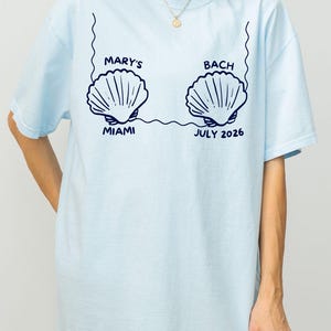 Puede incluir: Camiseta azul claro con dise&ntilde;o de conchas marinas. El texto "MARY'S BACH MIAMI JULIO 2026" es azul marino. El dise&ntilde;o incluye dos ilustraciones de conchas conectadas por una l&iacute;nea.