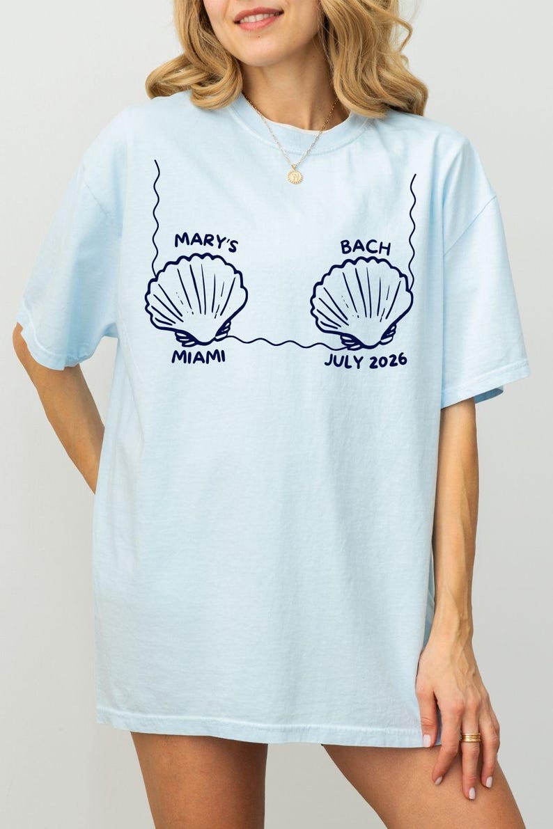 Puede incluir: Camiseta azul claro con dise&ntilde;o de conchas marinas. El texto "MARY'S BACH MIAMI JULIO 2026" es azul marino. El dise&ntilde;o incluye dos ilustraciones de conchas conectadas por una l&iacute;nea.