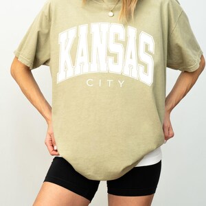 Peut inclure: Un t-shirt &agrave; manches courtes kaki avec le texte blanc KANSAS CITY &eacute;crit dans une police universitaire sur la poitrine. Le t-shirt pr&eacute;sente une encolure ronde et une coupe d&eacute;contract&eacute;e, associ&eacute; &agrave; un short de sport noir visible &agrave; l'ourlet.
