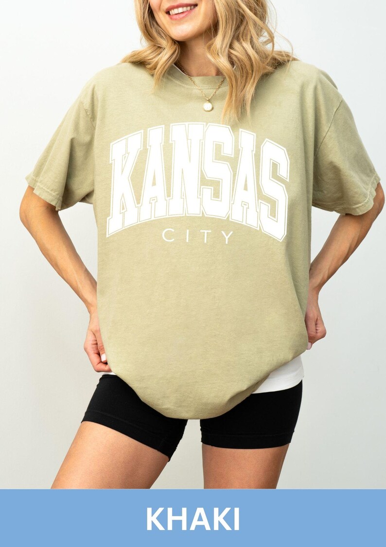Peut inclure: Un t-shirt &agrave; manches courtes kaki avec le texte blanc KANSAS CITY &eacute;crit dans une police universitaire sur la poitrine. Le t-shirt pr&eacute;sente une encolure ronde et une coupe d&eacute;contract&eacute;e, associ&eacute; &agrave; un short de sport noir visible &agrave; l'ourlet.