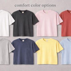 Puede incluir: Ocho camisetas de manga corta en varios colores: blanco, negro, rosa, rojo, gris, azul marino, amarillo y azul claro. El texto "comfort color options" se muestra en la parte superior.