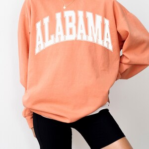 May include: A terracotta-colored crewneck sweatshirt featuring the word ALABAMA printed in a large, white, arched collegiate-style font across the chest. The long-sleeved garment has a relaxed fit and is paired with black athletic shorts.