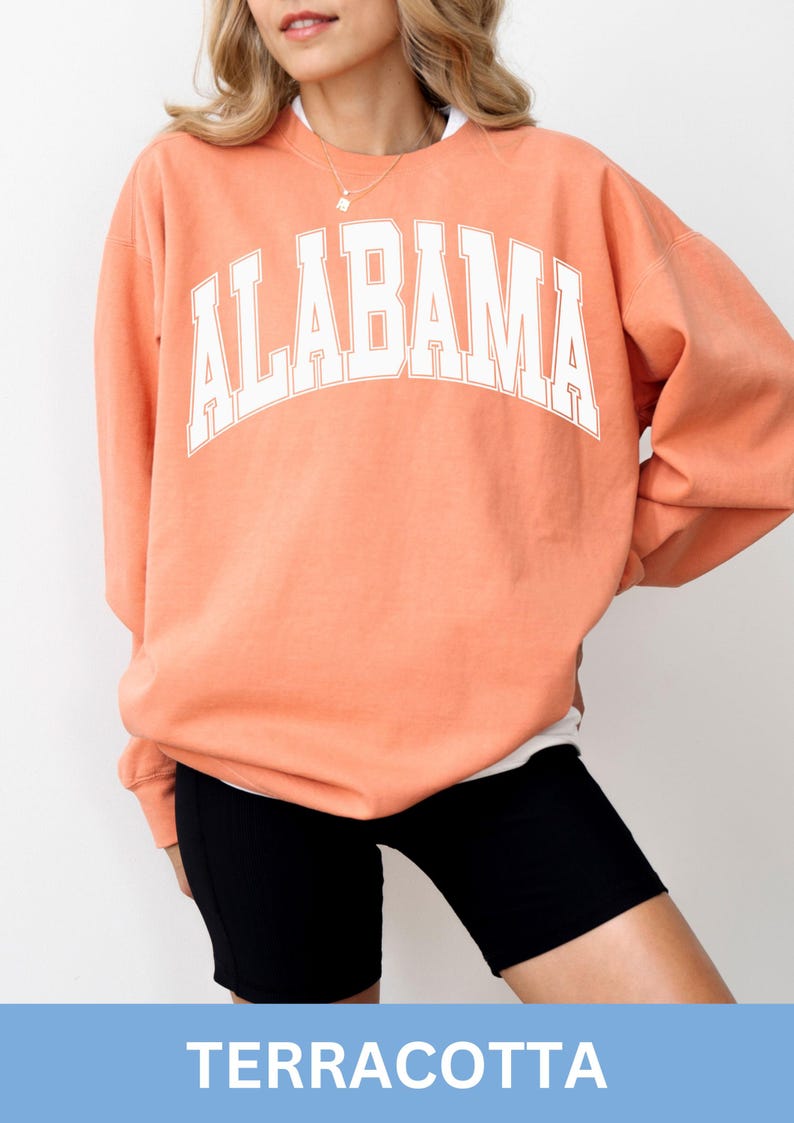 May include: A terracotta-colored crewneck sweatshirt featuring the word ALABAMA printed in a large, white, arched collegiate-style font across the chest. The long-sleeved garment has a relaxed fit and is paired with black athletic shorts.