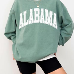May include: A sage green crewneck sweatshirt featuring the word ALABAMA printed in large white block letters across the chest. The garment has long sleeves, a ribbed neckline, and is layered over a white shirt, paired with black shorts.