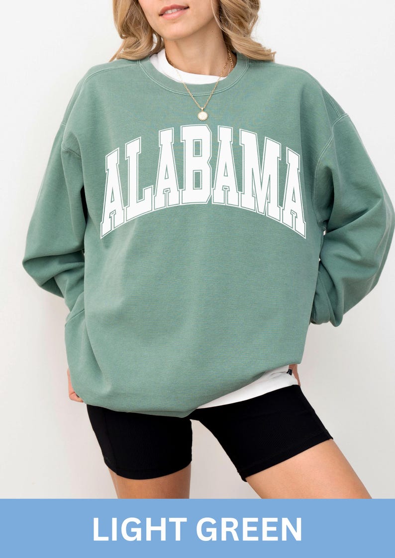 May include: A sage green crewneck sweatshirt featuring the word ALABAMA printed in large white block letters across the chest. The garment has long sleeves, a ribbed neckline, and is layered over a white shirt, paired with black shorts.